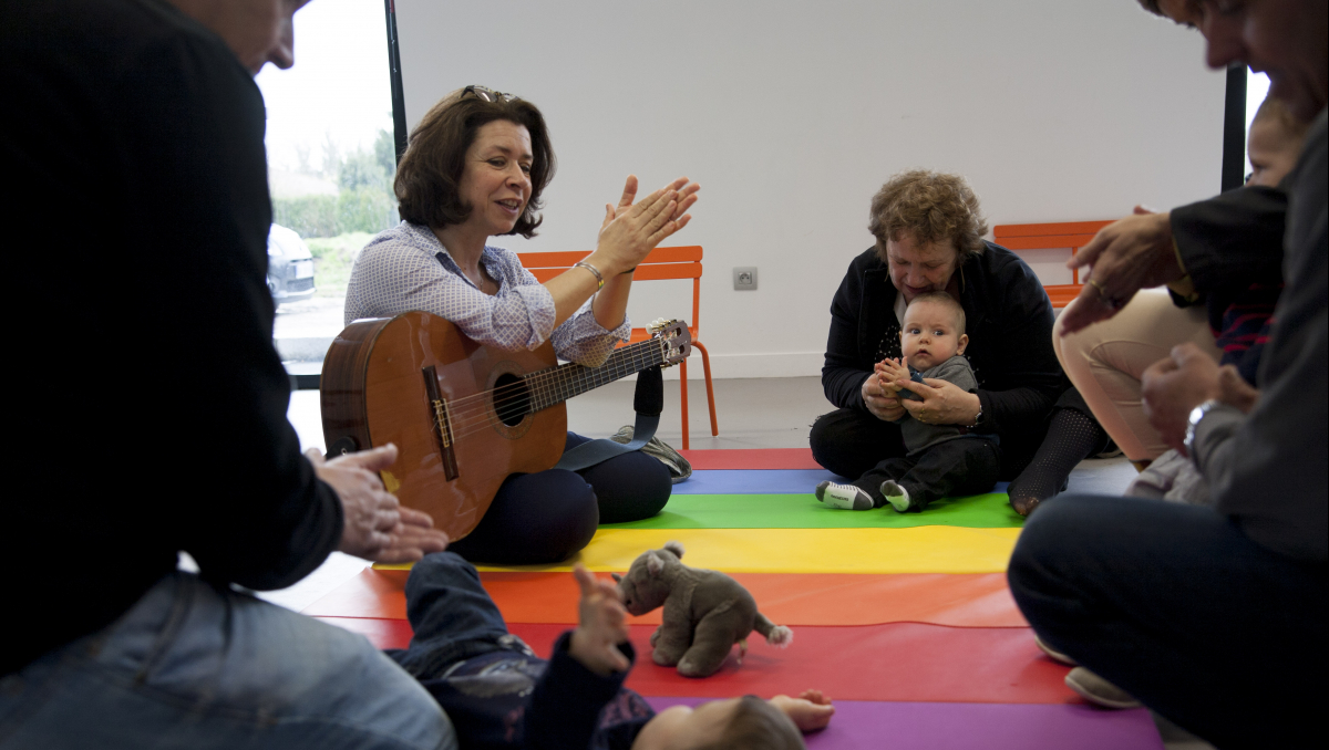 Une dame avec une guitare regarde un bébé sur un tapis